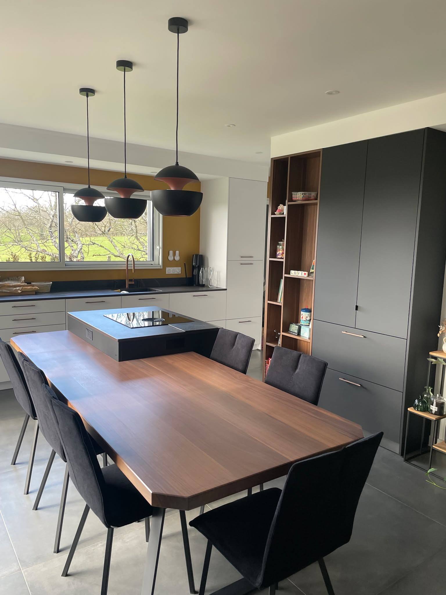 Table de salle à manger en noyer et chaises noires devant la cuisine, suspensions noires et mur bleu nuit, maison contemporaine